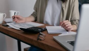 A woman manages finances at home, using a laptop and calculator on a wooden desk.